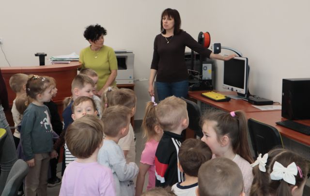 Na zdjęciu znajduje się grupa przedszkolaków w sali komputerowej, przy jednym z komputerów stoi kobieta i łapie ręką monitor, po lewej stronie stoi druga kobieta i biurko.