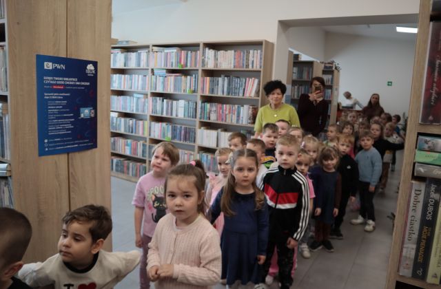 Na zdjęciu znajduje się grupa przedszkolaków, trzymają się za ręce i idą w parach między półkami z książkami w bibliotece, razem z nimi idą trzy kobiety.