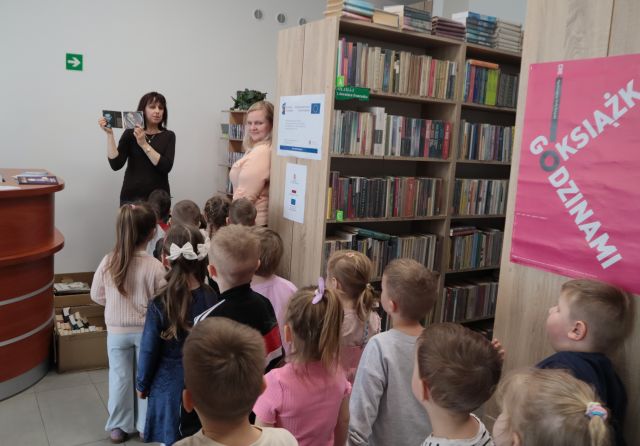 Na zdjęciu znajdują się przedszkolaki stojące w parach, jeden za drugim, przed nimi stoi kobieta i pokazuje im audiobook, a obok stoi inna kobieta. Po prawej stronie znajdują się półki z książkami.