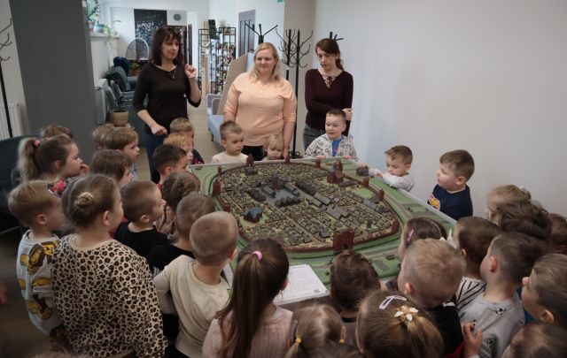 Na zdjęciu znajdują się dzieci oraz trzy kobiety dookoła makiety średniowiecznego miasta na holu biblioteki.