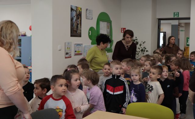 Na zdjęciu znajduje się grupa przedszkolaków oraz cztery kobiety w bibliotece, po lewej stronie na ścianie wiszą zdjęcia, gablota w kształcie żółwia.