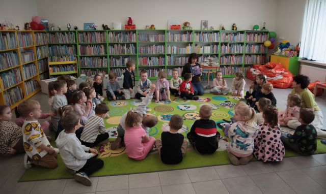 Na zdjęciu znajduje się czterdzieści przedszkolaków na dywanie wraz z dwoma kobietami, jedna z nich trzyma w ręku książkę. W tle znajdują się półki z książkami.