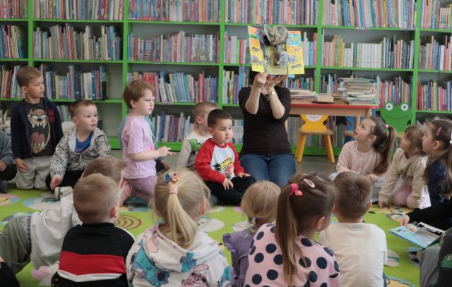 Na zdjęciu znajdują się przedszkolaki na dywanie, na środku siedzi kobieta i pokazuje im książkę, z której wystaje słoń. W tle znajduje się stolik z książkami oraz półki z książkami.