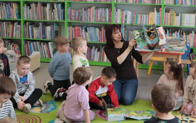 Na zdjęciu znajdują się dzieci na dywanie wraz z kobietą, która pokazuje im książki 3D. W tle znajdują się półki z książkami.
