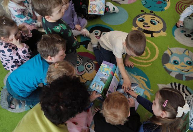 Na zdjęciu znajdują się dzieci na dywanie i wspólnie przeglądają książkę. Zdjęcie zostało wykonane od góry.