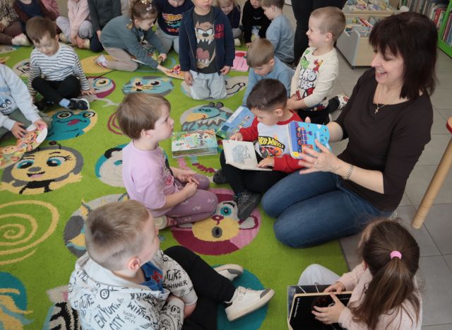 Na zdjęciu znajdują się dzieci na dywanie, które przeglądają książki. Po prawej stronie siedzi kobieta i pokazuje dziewczynce na przeciwko książkę.