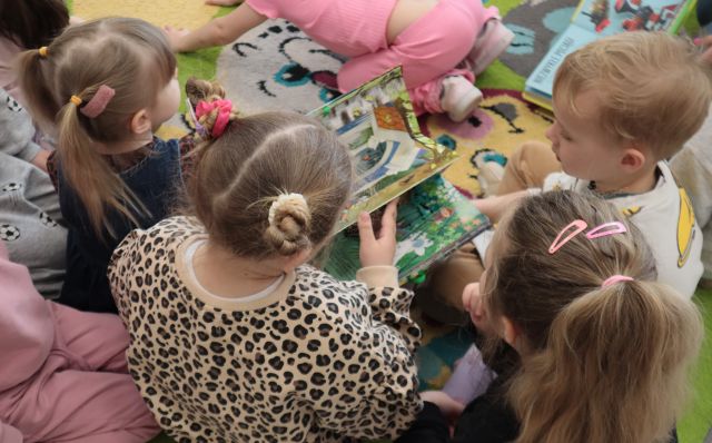 Na zdjęciu znajduje się grupa przedszkolaków oglądająca książkę na dywanie. Zdjęcie zostało wykonane od góry.