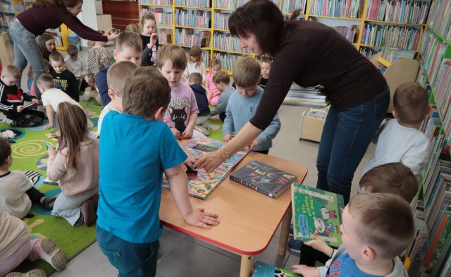 Na zdjęciu znajduje się kobieta nachylająca się przy stoliku z książkami wraz z grupą dzieci. W tle znajdują się przedszkolaki na dywanie i przeglądają książki oraz półki z książkami.