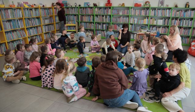 Na zdjęciu znajduje się czterdziestu przedszkolaków ora pięć kobiet na dywanie, kobieta siedząca na środku trzyma w ręce kartę i pokazuje dzieciom. W tle znajdują się półki z książkami.