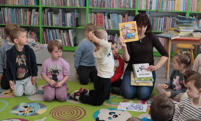 Na zdjęciu znajduje się kobieta trzymająca w ręce książkę i kilka książek na kolanach, dookoła niej siedzą przedszkolaki, a w tle znajdują się półki z książkami.