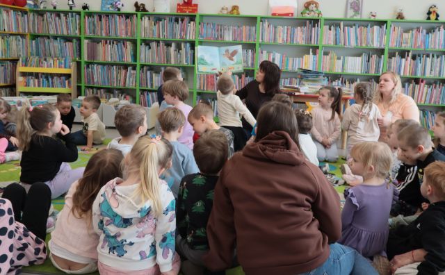Na zdjęciu znajdują się przedszkolaki oraz dwie kobiety na dywanie, na przeciwko nich siedzi kobieta i pokazuje im ilustrację w książce. W tle znajdują się półki z książkami.