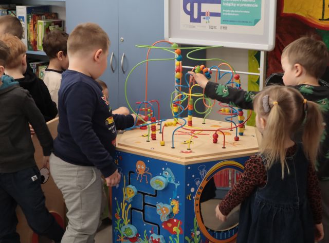 Na zdjęciu znajdują się przedszkolaki bawiące się przy wielofunkcyjnym stoliku edukacyjnym. W tle znajdują się szafki.