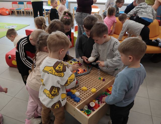 Na zdjęciu znajdują się dzieci bawiące się przy stoliku edukacyjnym do majsterkowania. W tle znajdują się bawiące się przedszkolaki.