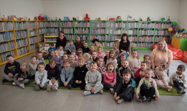 Na zdjęciu znajduje się czterdzieści przedszkolaków wraz z pięcioma kobietami na dywanie i uśmiechają się do zdjęcia. W tle znajdują się półki z książkami.