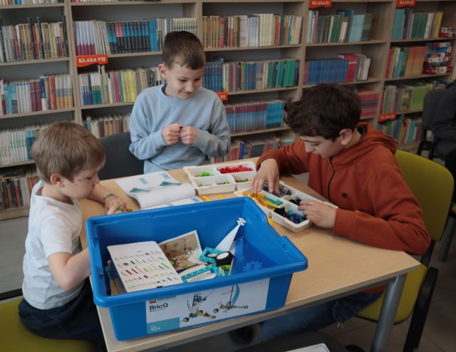 Na zdjęciu znajduje się trójka chłopców przy stoliku, przy którym składają klocki LEGO. W tle znajdują się półki z książkami.