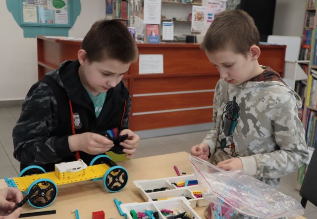 Na zdjęciu znajduje się dwójka chłopców, którzy wspólnie układają klocki LEGO znajdujące się na stoliku przed nimi. W tle znajduje się biurko.