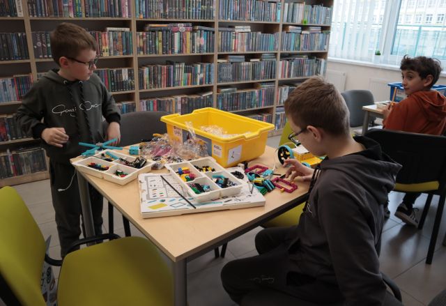 Na zdjęciu znajduje się dwójka chłopców przy stoliku i układają konstrukcje z klocków LEGO. Po prawej stronie przy sąsiednim stoliku siedzi chłopiec. W tle znajdują się półki z książkami.