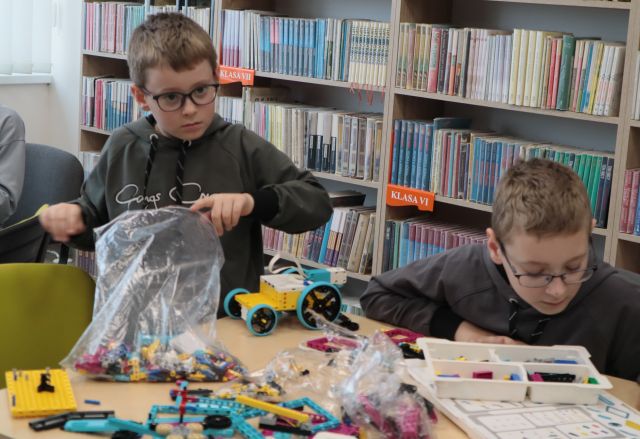 Na zdjęciu znajduje się dwójka chłopców przy stoliku, jeden z nich układa auto z klocków, a drugi szuka odpowiedniego klocka w torbie z LEGO. W tle znajdują się półki z książkami.