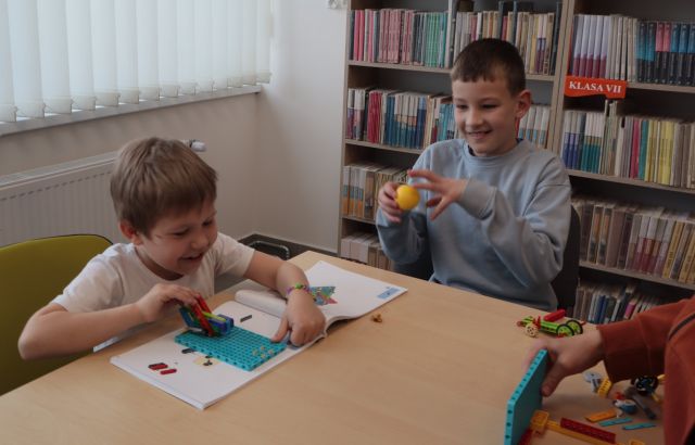 Na zdjęciu znajduje się dwóch chłopców przy stoliku, jeden z nich trzyma w ręce małą plastikową piłeczkę, a drugi ma przed sobą instrukcję i konstrukcję z klocków LEGO, na przeciwko niego leży parę klocków. W tle po lewej stronie znajdują się okna, a po prawej półki z książkami.