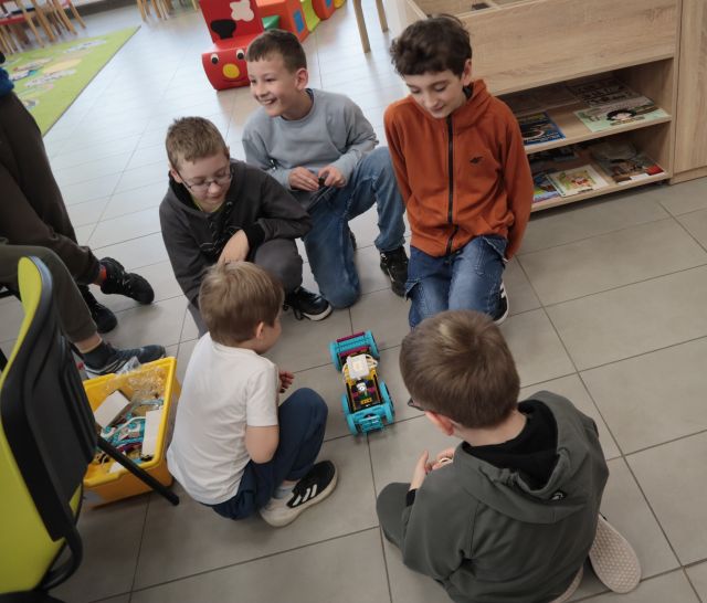 Na zdjęciu znajduje się pięciu chłopców na podłodze, po środku nich stoi samochód zbudowany z klocków LEGO. Po lewej stronie stoi krzesło, a za chłopcem, po prawej stronie stoi szafka.