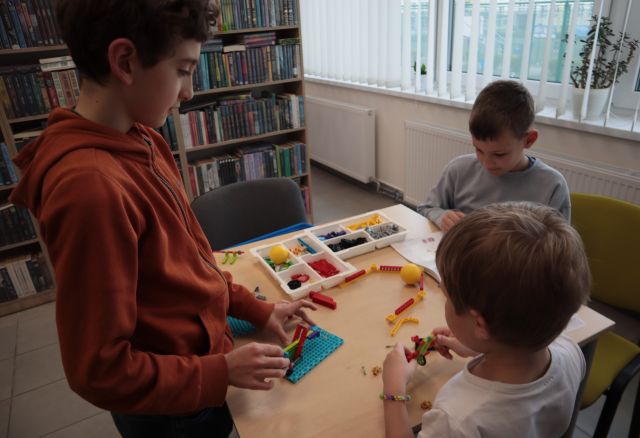 Na zdjęciu znajduje się trójka chłopców przy stoliku, przy którym układają klocki LEGO. W tle znajdują się półki z książkami i okna po prawej stronie.