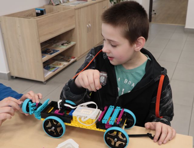 Na zdjęciu znajduje się chłopiec przy stoliku i ma przed sobą auto zbudowane z klocków LEGO. W tle znajdują się szafki.