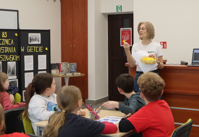 Na zdjęciu znajduje się kobieta, która trzyma w dłoni żonkila z papieru i pokazuje siedzącym przed nią dzieciom. Warsztaty odbywają się w sali komputerowej.