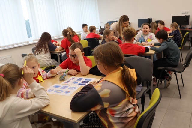 Na zdjęciu znajdują się uczniowie przy pięciu kwadratowych stolików i rozwiązują grupowo zadania, przy jednym z nich nachyla się kobieta. Warsztaty odbywają się w sali komputerowej.