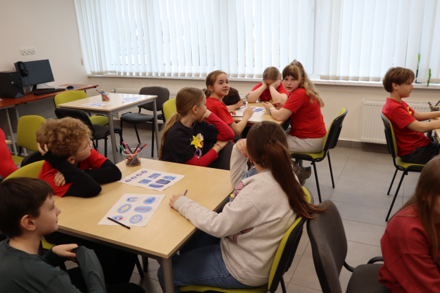 Na zdjęciu znajdują się uczniowie przy stolikach i rozwiązują zadania, jeden stolik stoi pusty. Warsztaty odbywają się w sali komputerowej i w tle znajdują się okna.