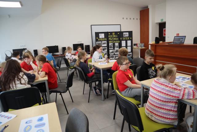 Na zdjęciu znajdują się uczniowie przy pięciu stolikach i rozwiązują zadania, w tle znajduje się tablica nawiązująca do rocznicy wybuchu powstania w getcie warszawskim. Warsztaty odbywają się w sali komputerowej.