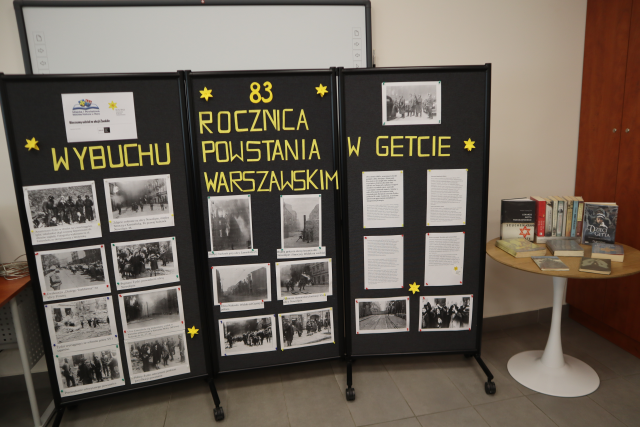 Na zdjęciu znajduje się czarna tablica ze zdjęciami i opisami upamiętniająca 83 rocznice wybuchu powstania w getcie warszawskim, po prawej stronie stoi stoliczek z książkami. 