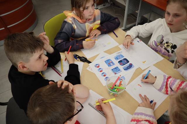 Na zdjęciu znajduje się piątka uczniów przy stoliku i rozwiązują leżące na stoliku zadania, w dłoni trzymają kredki. Zdjęcie zostało wykonane od góry.