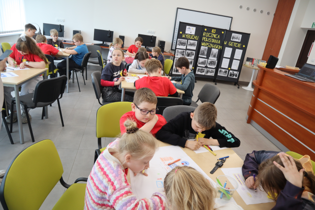 Na zdjęciu znajdują się uczniowie przy stolikach w sali komputerowej i rozwiązują zadania. W tle po prawej znajduje się tablica upamiętniająca 83 rocznice wybuchu powstania w getcie warszawskim.