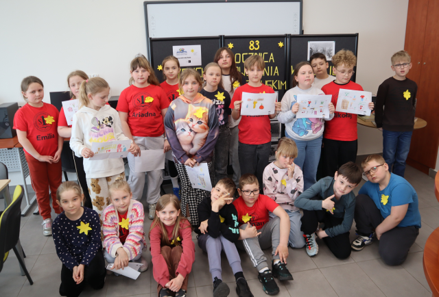 Na zdjęciu znajduje się dwudziestu jeden uczniów, część ma w ręce kartki i przyklejone na koszulce żółte żonkile. Uczniowie stoją do zdjęcia na tle czarnej tablicy upamiętniającej 83 rocznice wybuchu powstania w getcie warszawskim. 