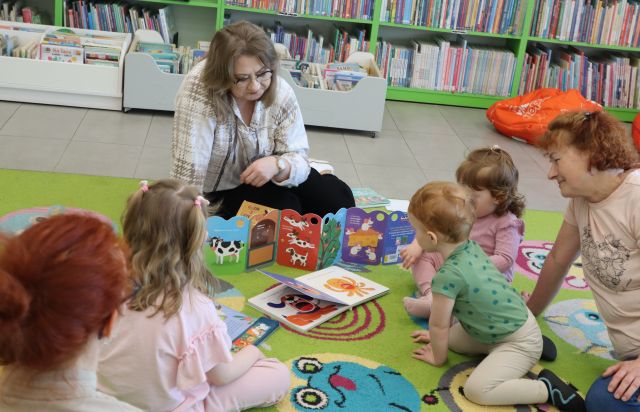 Na zdjęciu znajdują się maluchy wraz z rodzicami oraz kobieta na środku, która pokazuje im rozkładaną książeczkę. Uczestnicy siedzą na dywanie, a w tle znajdują się półki z książkami.