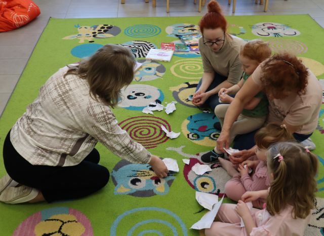 Na zdjęciu znajduje się trójka dorosłych kobiet oraz trójka dzieci i rwą kawałek kartki na mniejsze części. Zdjęcie zostało wykonane od góry.