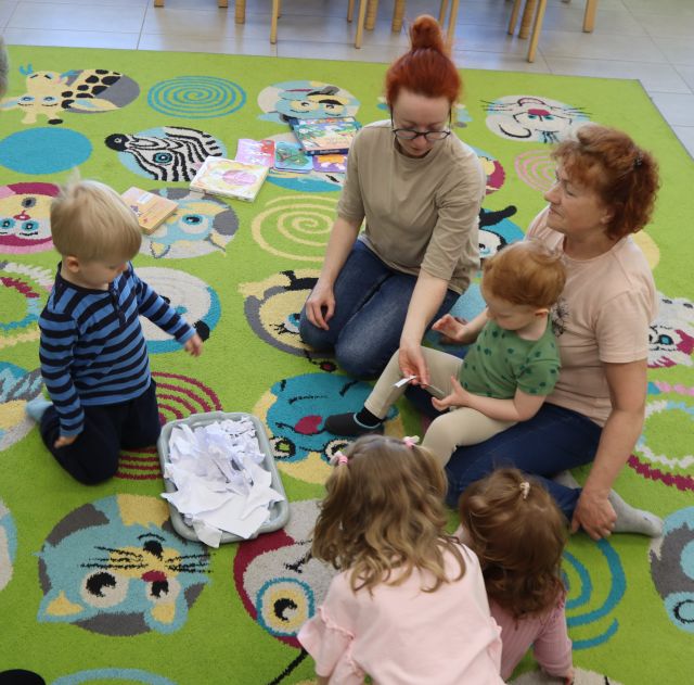 Na zdjęciu znajduje się czwórka maluchów oraz dójka dorosłych na dywanie, na środku na tacce leżą porwane kawałki papieru.