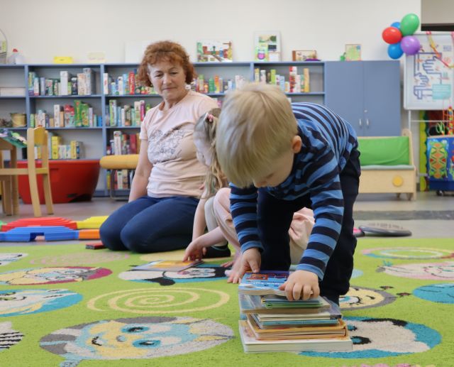 Na zdjęciu znajduje się chłopiec nachylony nad książką na dywanie, w tle znajduje się kobieta i dziewczynka schowana za chłopcem. W tle znajdują się półki z książkami.