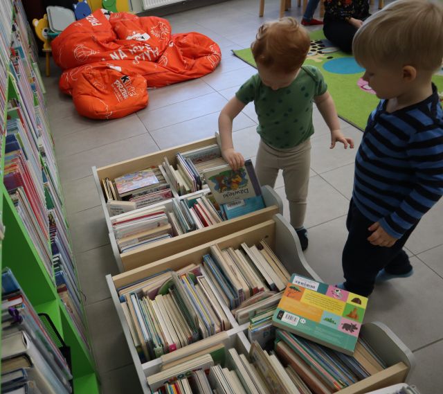 Na zdjęciu znajduje się dwóch chłopców przy wózeczkach z książkami, w tle znajdują się czerwone pufy.
