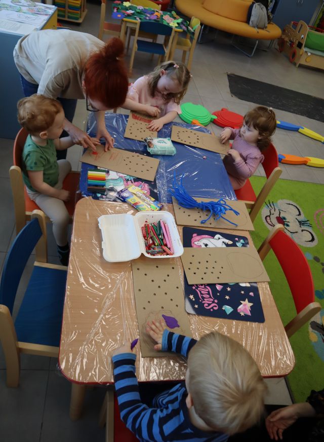 Na zdjęciu znajduje się czwórka dzieci przy złączonych stoliczkach i ozdabiają kartonowe doniczki plasteliną. Na stoliku znajdują się również kredki i druciki, obok nachyla się kobieta. 