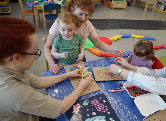 Na zdjęciu znajdują się dwie kobiety oraz dwójka dzieci przy stoliku, jedna kobieta trzyma chłopca na kolanach i wspólnie przeciągają druciki kreatywne przez dziurki. W tle znajduje się rozłożona na podłodze ścieżka sensoryczna.