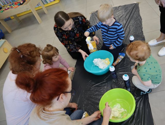 Na zdjęciu znajduje się trójka dorosłych oraz trójka dzieci i robią w dwóch miskach masę. Na podłodze jest rozłożony kawałek czarnej folii.