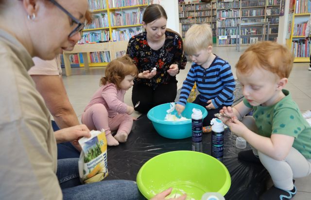 Na zdjęciu znajduje się trójka dzieci oraz dwie kobiety na podłodze, na rozciągniętym kawałku czarnej folii i wspólnie robią masę w miskach. W tle znajdują się półki z książkami.