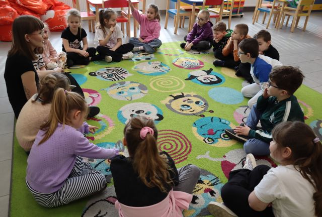 Na zdjęciu znajdują się przedszkolaki siedzące na dywanie wraz z kobietą. W tle znajdują się stoliczki z krzesłami.