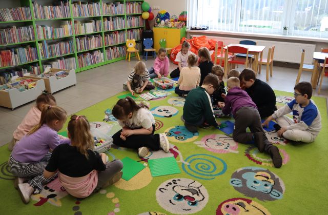 Na zdjęciu znajduje się piętnastu przedszkolaków, które w trzech grupach na dywanie układają klocki LEGO. W tle znajdują się półki z książkami, pufy i stoliczki.