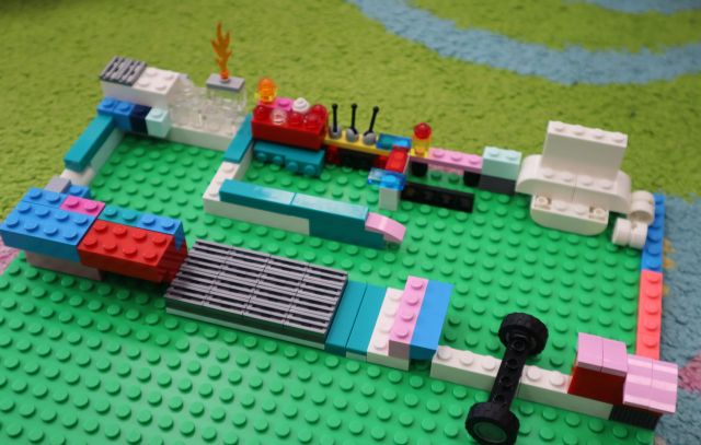 Na zdjęciu znajduję się pomieszczenie biblioteczne zbudowane z klocków LEGO na zielonej podkładce. Praca znajduje się na dywanie.