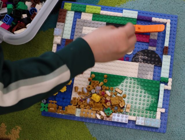 Na zdjęciu znajduje się ręka chłopca, który dokłada klocki do swojej budowli z klocków LEGO, która znajduje się na niebieskiej podkładce na dywanie. W lewym górnym rogu znajduje się fragment pudełka z klockami.