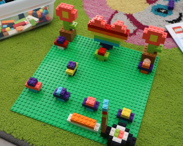 Na zdjęciu znajdują się zbudowane z klocków LEGO serduszka, kwiatki, sushi na zielonej podkładce, która leży na dywanie. W prawym górnym rogu znajduje się pudełko z klockami.