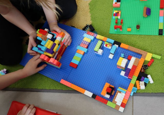 Na zdjęciu znajduje się zbudowana z klocków LEGO biblioteka na dwóch niebieskich podkładkach, z lewej strony budowla trzymana jest przez dziewczynki, które dokładają kolejne elementy. Praca znajduje się na dywanie, a w prawym górnym rogu leży zielona podkładka.