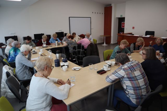 Na zdjęciu znajduje się około dwudziestu kobiet siedzących przy złączonych stołach w literkę C i rozwiązują zadania przy kawie. Warsztaty odbywają się w sali komputerowej.